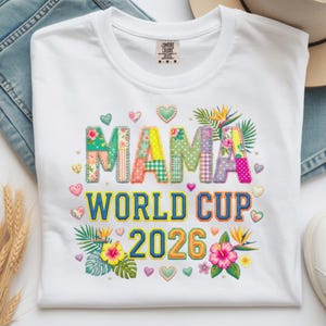 Peut inclure: T-shirt blanc avec l'inscription "MAMA WORLD CUP 2026" en lettres et chiffres patchwork colorés. Le motif comprend des accents floraux et en forme de cœur. Le t-shirt est en matière douce.