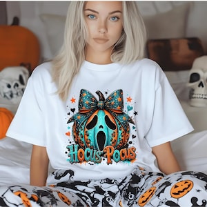 Peut inclure: Un t-shirt blanc à manches courtes avec un motif de citrouille bleu sarcelle et orange orné d'un grand nœud et d'une expression faciale criante. Le texte Hocus Pocus est imprimé sous la citrouille dans une police bleue et orange stylisée.