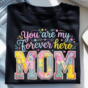 Peut inclure: T-shirt noir avec l'inscription "You are my forever hero MOM" en lettres brodées colorées. Le mot "MOM" est orné de motifs patchwork et d'éléments floraux, parfait pour la fête des mères.