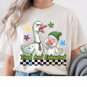 Peut inclure: T-shirt crème avec une illustration de dessin animé de deux oies. L'une porte une cravate et a une expression de colère, tandis que l'autre porte un bonnet vert et un nœud papillon rose. Le texte "Tired And Exhausted" est imprimé en dessous.