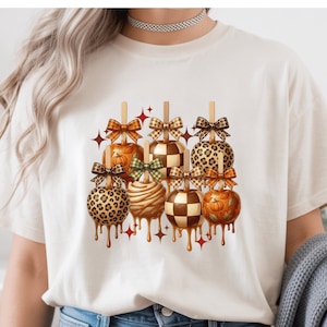 May include: A cream-colored t-shirt featuring a graphic print of seven caramel apples on sticks. The apples display various patterns including leopard print, checkered designs, and pumpkin motifs, each topped with a decorative bow and red star accents.