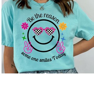 Peut inclure: T-shirt bleu clair avec un motif de smiley portant des lunettes de soleil en forme de cœur à carreaux. Le t-shirt porte l'inscription "Be the reason some one smiles Today" dans un motif circulaire avec des fleurs et des signes de paix.