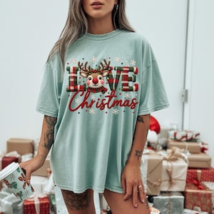 Peut inclure: Un t-shirt vert clair avec un motif portant l'inscription LOVE Christmas. La lettre O est remplacée par une tête de renne au nez rouge avec des bois. Les lettres L, V et E présentent un motif à carreaux rouge et vert. Des flocons entourent le texte.