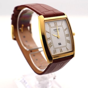 May include: A gold-toned Seiko Quartz watch with a rectangular white face and Roman numeral hour markers. The watch has a brown leather band. The watch face displays the Seiko logo and the word "QUARTZ".