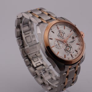 May include: A stainless steel wristwatch with a white face and rose gold accents. The watch features a chronograph design with three sub-dials and a date display. The watch band is a combination of stainless steel and rose gold links.