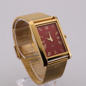 May include: Gold-toned rectangular wristwatch with a burgundy face and gold hands and Roman numeral hour markers. The watch has a mesh band and a small crown. The watch is displayed on a clear stand.