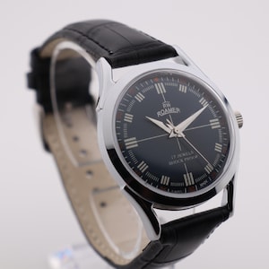 May include: A vintage Roamer watch with a black dial and silver hands. The watch has a black leather strap and a stainless steel case. The dial features the brand name and the words "17 Jewels Shockproof".