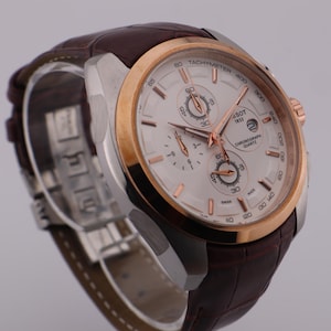 May include: A Tissot chronograph watch with a white face, gold bezel, and brown leather strap. The watch features multiple subdials and rose gold hands and accents. The watch is displayed on a clear stand.