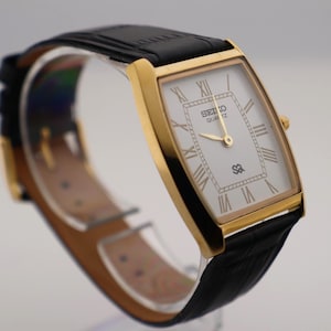 May include: A gold-toned rectangular Seiko Quartz watch with a white face and black leather strap. The watch face features Roman numerals and gold hands. The watch is displayed against a neutral background.