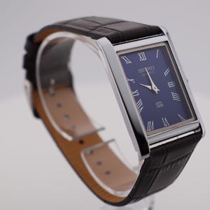 May include: A rectangular Seiko Quartz watch with a dark blue face and silver frame. The watch features Roman numeral hour markers and a black leather strap. The watch is displayed on a clear stand.