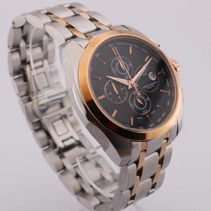 May include: A silver and gold-tone wristwatch with a black face. The watch features three sub-dials and a date window. The watch band is silver and gold-tone. The watch has a gold-tone bezel and rose gold-tone hands and markers.