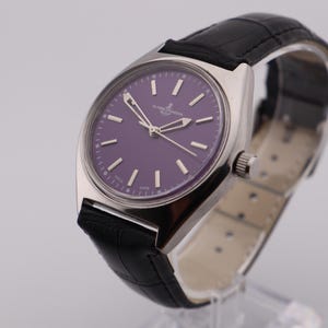May include: A silver wristwatch with a purple face and a black leather strap. The watch features silver hands and markers, and the brand name is visible on the face. The strap is textured.