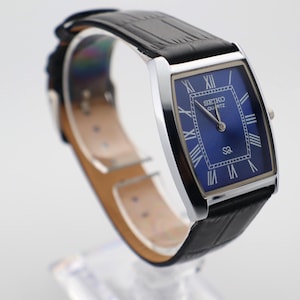 Puede incluir: Un reloj Seiko Quartz rectangular con una correa de cuero negro. La esfera es azul oscuro con números romanos y agujas plateadas. El reloj se muestra sobre un soporte transparente.