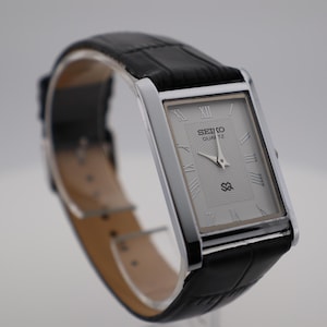May include: A rectangular Seiko Quartz watch with a silver face and black leather strap. The watch face features Roman numerals and the Seiko logo. The strap has a textured, crocodile-style pattern.