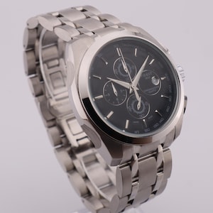 May include: A stainless steel wristwatch with a black dial. The watch features three sub-dials and luminous hands. The band is made of stainless steel. The watch is a chronograph style.