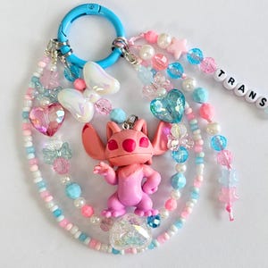 May include: A handmade keychain featuring a pink Stitch figurine, surrounded by pastel beads, iridescent butterfly charms, and heart-shaped accents. The keychain includes a blue clasp and a strand of beads spelling "TRANS PRIDE".
