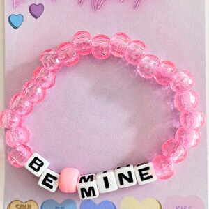 May include: A pink beaded bracelet spelling "BE MINE" with white letter beads. The bracelet sits on a pink card with "LunaKey Designs" and heart-shaped candies with phrases like "Soul Mate" and "Cutie".