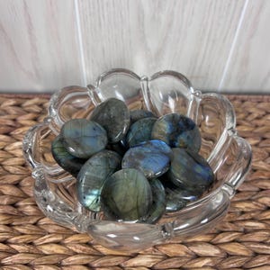 May include: A clear, scalloped glass bowl filled with polished labradorite stones. The stones display iridescent flashes of blue, green, and gold. The bowl sits on a woven, brown surface.
