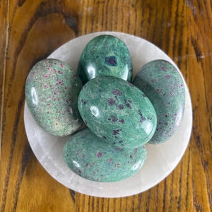 May include: Five polished green stones with red speckles rest in a white, round bowl. The stones have an oval shape and a glossy finish. The bowl is set on a wooden surface, creating a natural and earthy aesthetic.