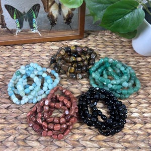 Puede incluir: Una variedad de pulseras de piedras preciosas en varios colores, incluyendo azul claro, marrón, verde, rojo-marrón y negro. Las pulseras están hechas de piedras pulidas y se presentan sobre una superficie tejida. La imagen también muestra una exhibición enmarcada de mariposas.