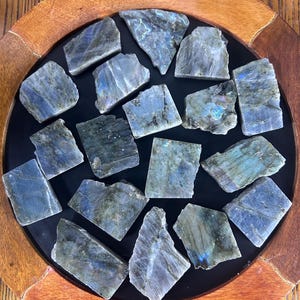 Puede incluir: Una colección de piedras de labradorita en bruto, que muestran una mezcla de tonos azules, grises e iridiscentes. Las piedras están dispuestas sobre una superficie negra dentro de una bandeja de madera con un borde marrón oscuro. Destacan las formas y colores naturales de las piedras.