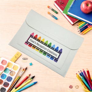 May include: A pale grey document wallet with a flap closure. It features a colourful crayon design with the text "Foxx's Reception Creations". Pencils, paintbrushes, and notebooks are scattered around the wallet.