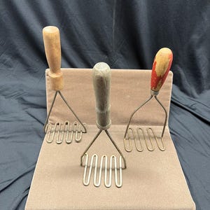 May include: Three vintage potato mashers with wooden handles and metal mashing heads. The handles vary in color, with one being light wood, one gray, and one red and yellow. They are displayed on a brown, rectangular stand.