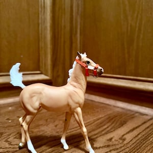 May include: A tan-colored toy horse figurine with a red halter. The horse has white markings on its legs and tail. The figurine is standing on a wooden surface.