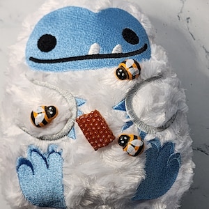 Pode incluir: Um yeti de pelúcia branco com um rosto azul e um sorriso cheio de dentes. O yeti está segurando um favo de mel e tem três abelhas amarelas e pretas nele.