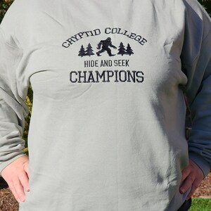May include: Sage green sweatshirt with the embroidered text "CRYPTID COLLEGE" above a Bigfoot graphic and the words "HIDE AND SEEK CHAMPIONS". The sweatshirt is a crew neck style.