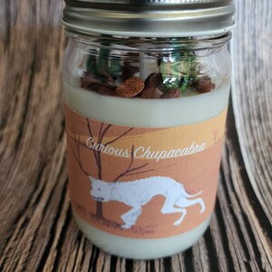 May include: A glass jar candle with a white label featuring a cartoon illustration of a white dog with the text "Curious Chupacabra". The candle has a light green wax with brown and green sprinkles.