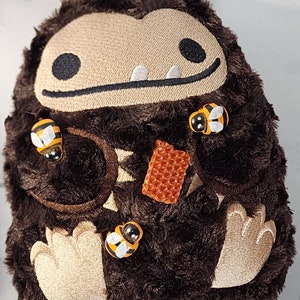 May include: A brown plush toy with a toothy grin and black eyes. The toy is covered in brown fur and has small, beige paws. There are four yellow and black bee-shaped buttons attached to the toy, and a small orange honeycomb-shaped button.