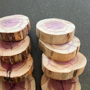 May include: A set of eight wooden coasters made from cedar wood. The coasters are round and have a natural wood finish with a reddish-brown hue. The coasters are stacked in two piles of four.