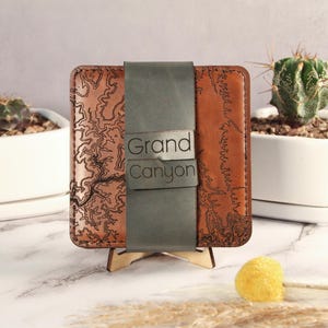 May include: A brown leather wallet with a detailed topographical map design. The wallet is secured with a gray leather strap that reads "Grand Canyon". The wallet is displayed on a wooden stand, with cacti in white pots in the background.