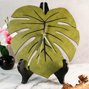 Puede incluir: Un reloj de pared decorativo con forma de hoja de monstera verde. El reloj tiene vetas negras y manecillas doradas. Se exhibe sobre un soporte negro, con una orquídea en maceta en el fondo. El diseño del reloj está inspirado en la naturaleza.