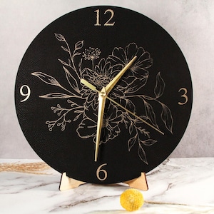 Puede incluir: Un reloj redondo negro con manecillas y números dorados. La esfera del reloj presenta un diseño floral dorado. El reloj está sostenido por un soporte de madera. El reloj tiene aproximadamente 25 cm de diámetro.