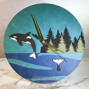 May include: Round wall clock with an orca whale design. The clock face has a blue and teal background, a black and white whale, a forest silhouette, and gold clock hands. The clock is mounted on a clear stand. The clock's diameter is approximately 30 cm.