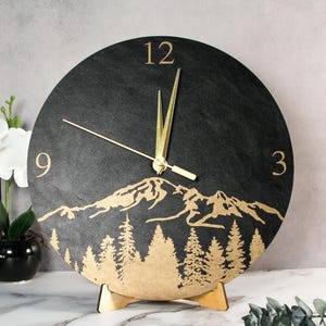 Puede incluir: Un reloj redondo negro con manecillas y números dorados. La esfera del reloj presenta un diseño de montaña y bosque en dorado. El reloj está sostenido por un soporte de madera. El reloj mide aproximadamente 25 cm de diámetro.