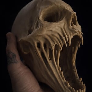 May include: A beige-coloured skull sculpture with an open mouth and sharp teeth. The skull is held in a hand against a black background, highlighting the detailed carving. The sculpture has a menacing, artistic appearance. The sculpture is made of a beige material.