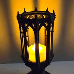 Könnte beinhalten: Eine schwarze Kerzenlaterne im gotischen Stil mit einer brennenden gelben Kerze im Inneren. Die Laterne hat ein verziertes, bogenförmiges Design und steht auf einem Sockel. Der Hintergrund ist warmgelb.