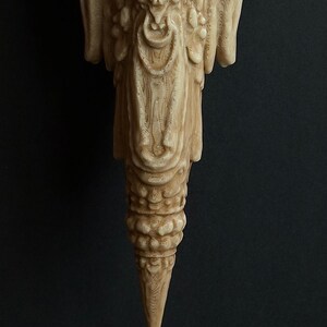 May include: A detailed, light-colored, carved wooden sculpture with a textured surface. The sculpture has an ornate design with a pointed bottom and a complex, layered structure. The background is a solid, dark color.