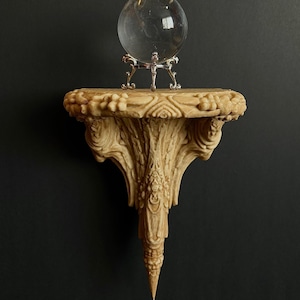May include: A clear glass crystal ball sits on a decorative, carved, beige-colored shelf. The shelf is mounted on a dark background. The shelf has ornate details and a pointed bottom.