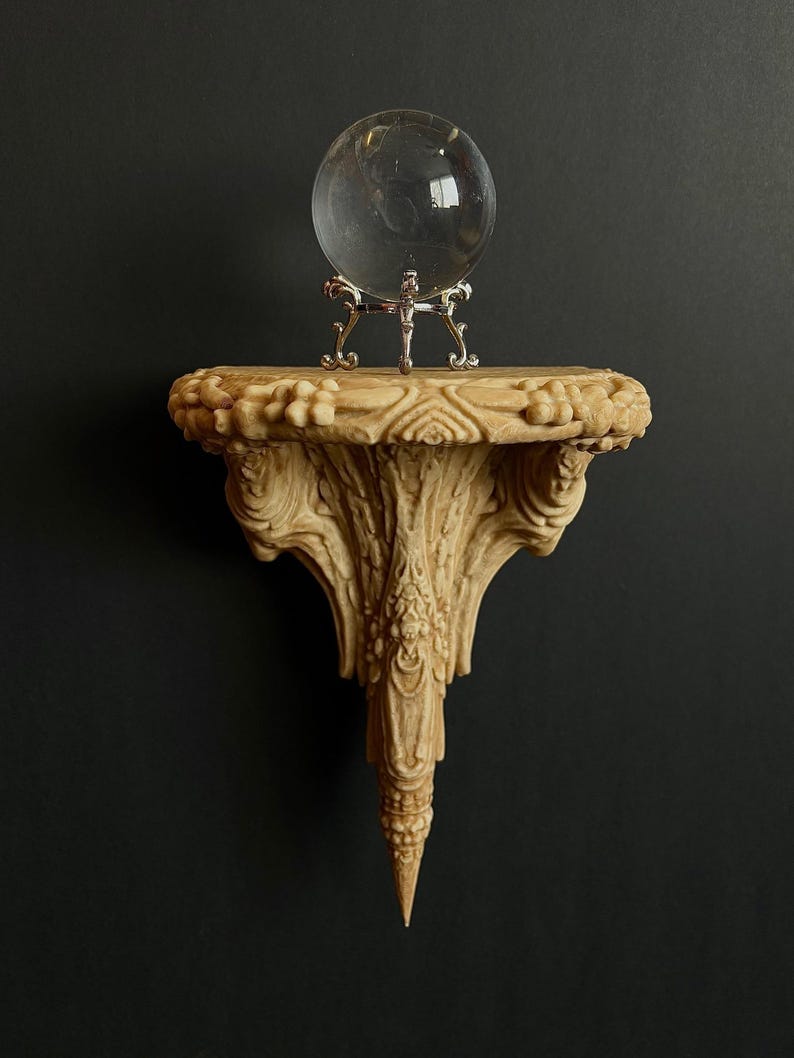 May include: A clear glass crystal ball sits on a decorative, carved, beige-colored shelf. The shelf is mounted on a dark background. The shelf has ornate details and a pointed bottom.