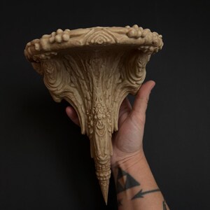 May include: Ornate beige wall shelf with a detailed, carved design. The shelf has a rounded top with a textured edge and a central, downward-tapering support. The shelf is held by a hand against a dark background.