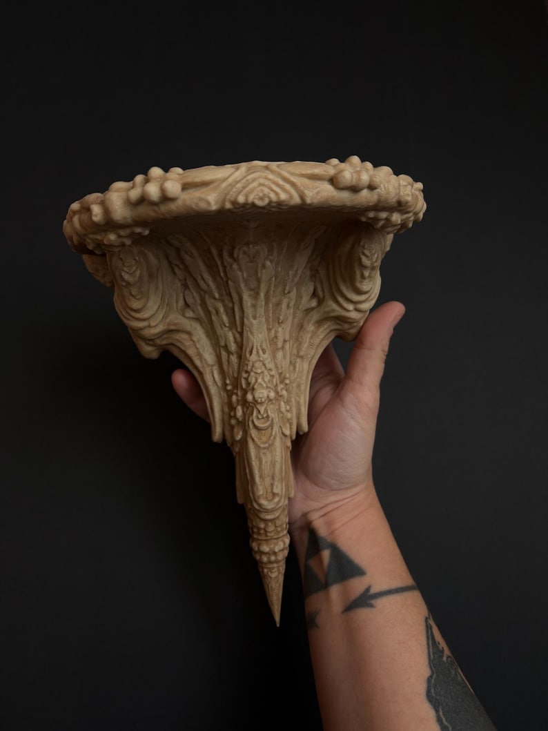 May include: Ornate beige wall shelf with a detailed, carved design. The shelf has a rounded top with a textured edge and a central, downward-tapering support. The shelf is held by a hand against a dark background.