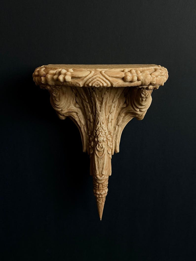 May include: A beige, ornate wall shelf with a detailed, carved design. The shelf has a rounded top with floral and geometric patterns, supported by a sculpted, tapering base. The shelf is against a dark background.