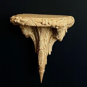 May include: Ornate, beige wall shelf with intricate carvings. The shelf has a rounded top with floral and scrollwork details. The support structure tapers to a point. The shelf is made of a light-colored material and is set against a black background.