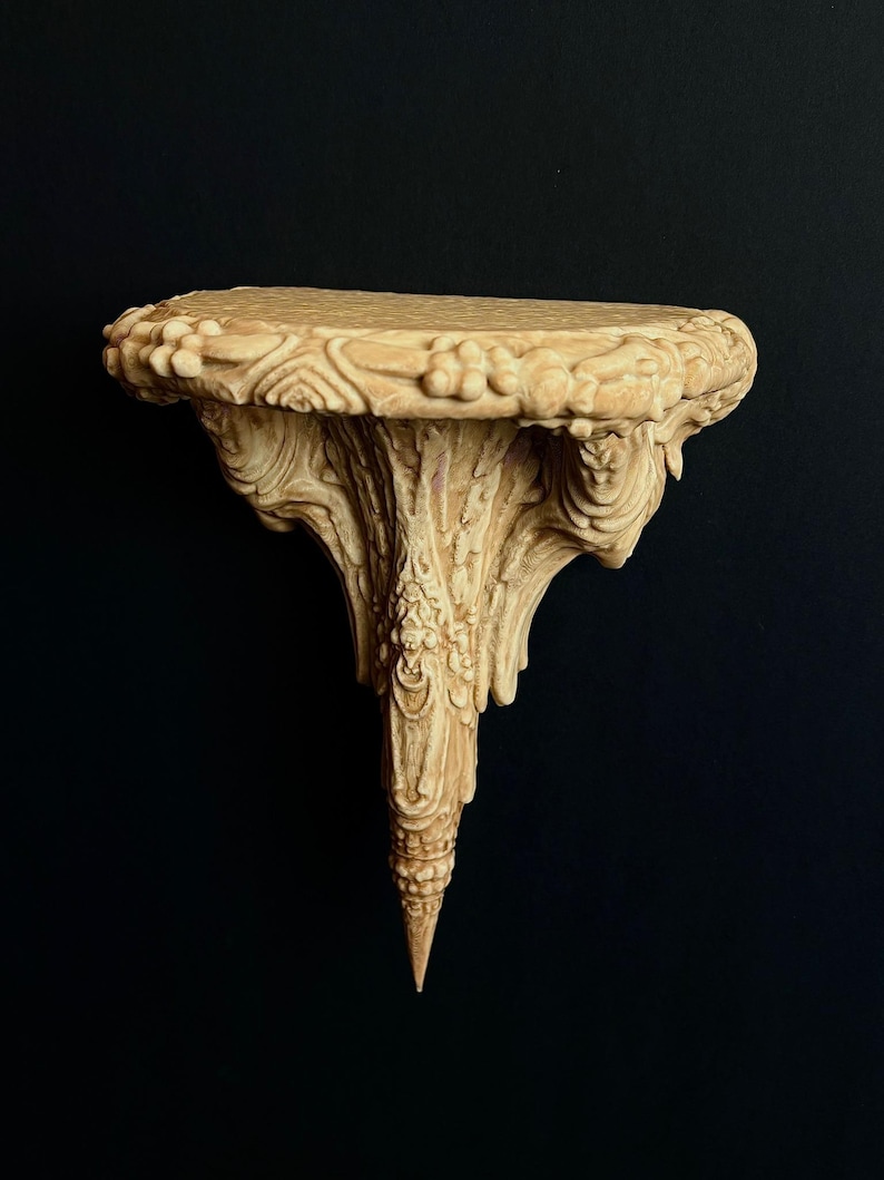 May include: Ornate, beige wall shelf with intricate carvings. The shelf has a rounded top with floral and scrollwork details. The support structure tapers to a point. The shelf is made of a light-colored material and is set against a black background.