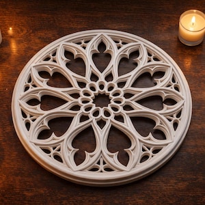 May include: A white, circular decorative piece with an intricate, symmetrical design, resembling a gothic rose window. Several lit candles in glass holders and a larger pillar candle surround the piece on a dark wooden surface.