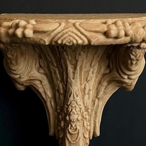 May include: Ornate, beige-colored wall shelf with detailed floral and leaf carvings. The shelf has a rounded top and a decorative, sculpted support. The design is reminiscent of classical architecture.
